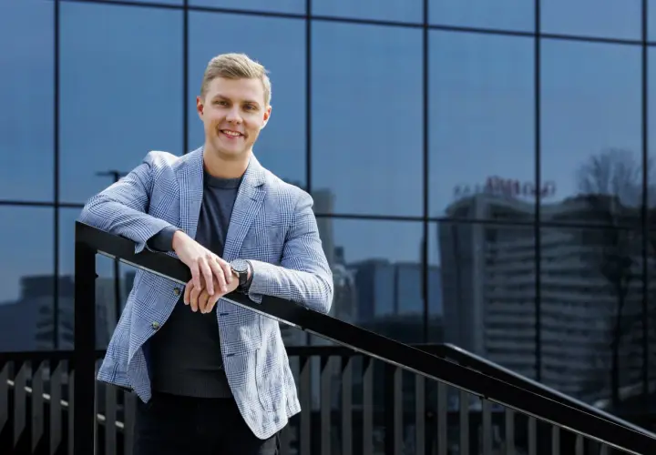 Uśmiechnięty mężczyzna w marynarce w kratę podczas sesji biznesowej w plenerze, oparty o balustradę na tle nowoczesnego budynku.