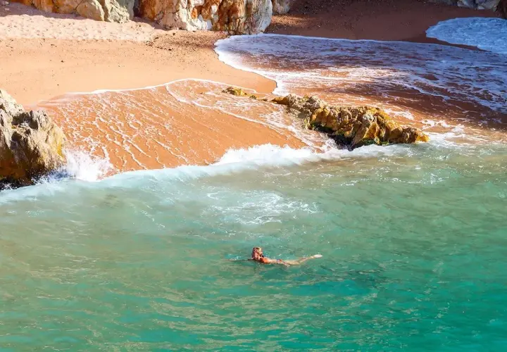Eine Frau schwimmt im türkisfarbenen Wasser vor einem goldenen costa brava strand, umgeben von Felsen.