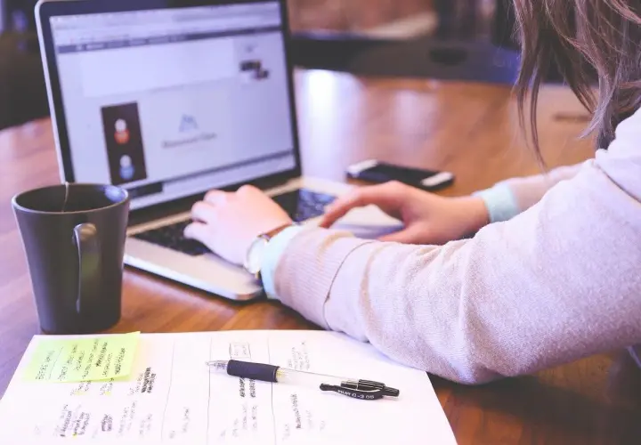 Kobieta pracuje przy laptopie. Na biurku leżą notatki, długopis i kubek. Dobre natężenie oświetlenia na stanowisku pracy sprzyja koncentracji.