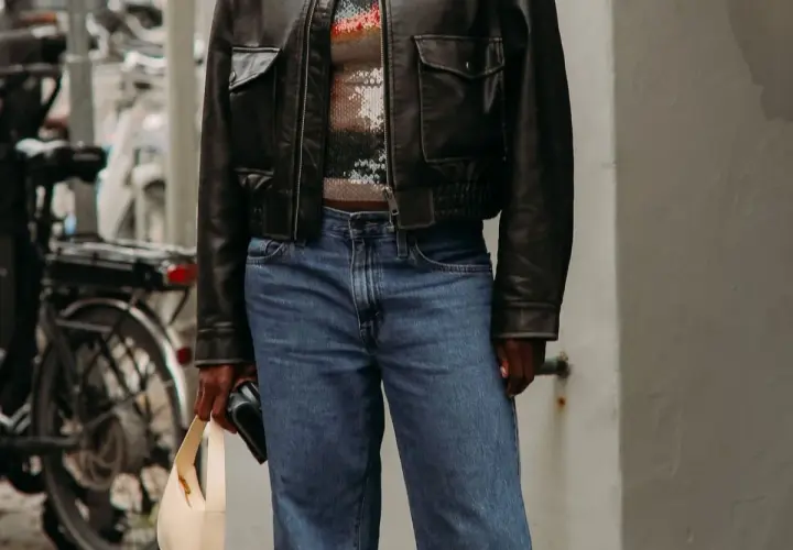 Personne portant une veste en cuir noire, un haut à paillettes et un jean bleu, tenant un sac beige.