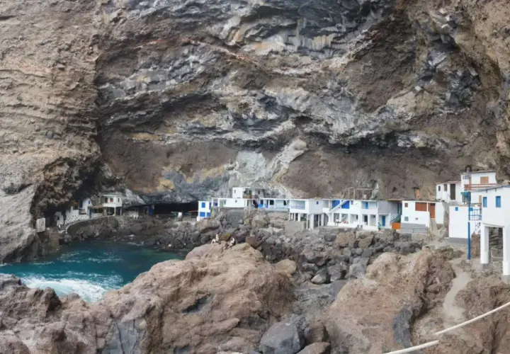 Casas blancas con detalles azules se asoman desde una cueva en los poris de Candelaria, La Palma. El mar turquesa rompe contra las rocas.