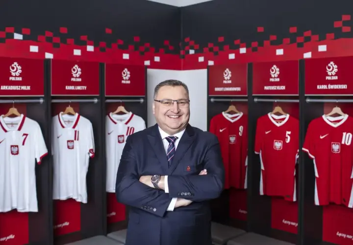 Uśmiechnięty mężczyzna w garniturze stoi przed szatnią reprezentacji Polski. Wiszą tam koszulki z nazwiskami Milika, Lewandowskiego i innych.