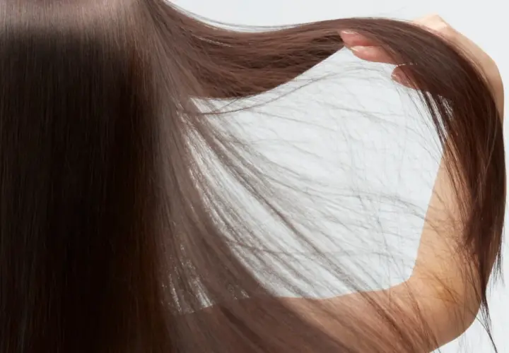 Mano acariciando cabello castaño, fino y escaso, que cae en cascada sobre un fondo blanco.