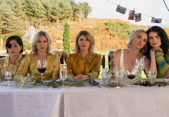 Fem kvinnor, kanske **bad sisters**, sitter vid ett dukat bord utomhus. En bär ögonlapp, en annan en glittrig gul klänning.