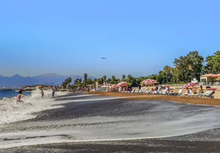 Ein sonniger Antalya Strand mit sanften Wellen, Liegestühlen und Palmen. Ein Flugzeug zieht seine Bahn am blauen Himmel.