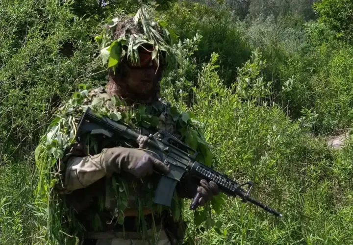 Żołnierz w pełnym kamuflażu, z liśćmi na głowie i ubraniu, trzyma karabin. Doskonałe maskowanie twarzy wojsko w zielonym otoczeniu.