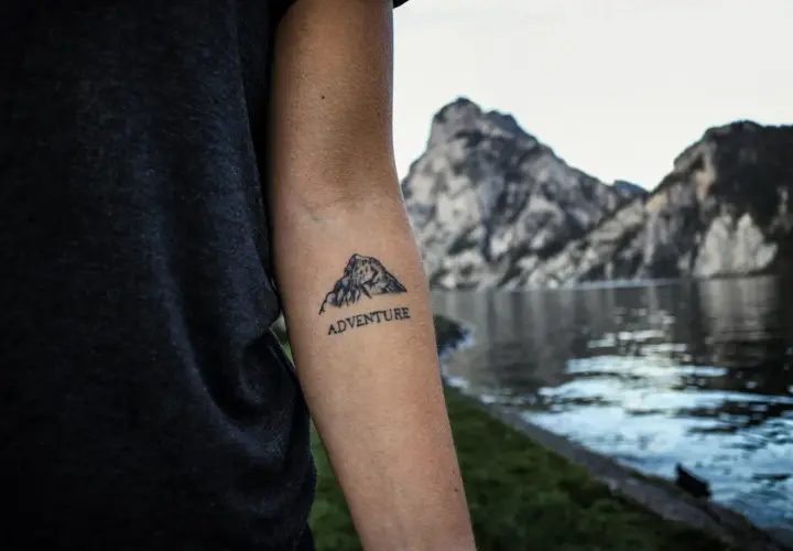 Tatuaż góry z napisem "ADVENTURE" na przedramieniu. W tle jezioro i skaliste szczyty.