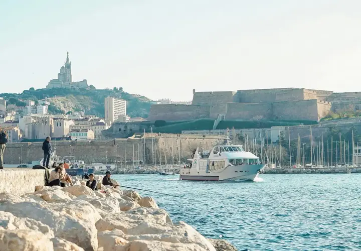 Widok na port w Marsylii z łodzią, fortecą i bazyliką na wzgórzu.