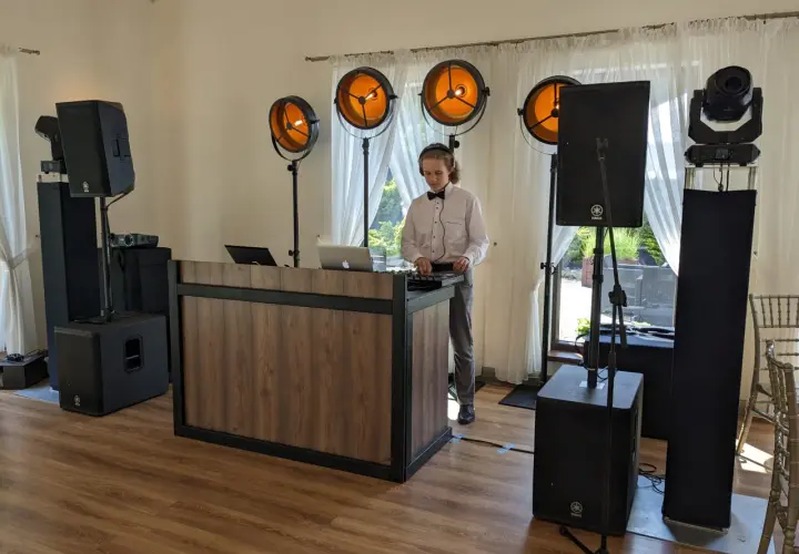 Młody DJ w muszce i białej koszuli obsługuje sprzęt muzyczny przy drewnianym stanowisku, otoczony głośnikami i oświetleniem.