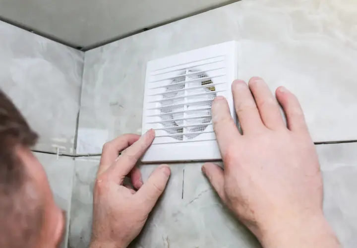 Installation d'un ventilateur pour l'aération de la salle de bain. Des mains ajustent l'appareil blanc sur un mur carrelé.