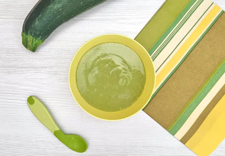 Un plato con puré de calabacín bebé, una cuchara verde y un calabacín entero sobre una mesa de madera.