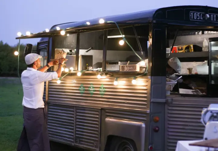 Un food truck illuminé de guirlandes lumineuses, parfait pour une idée food truck mariage. Un serveur sert un plat.