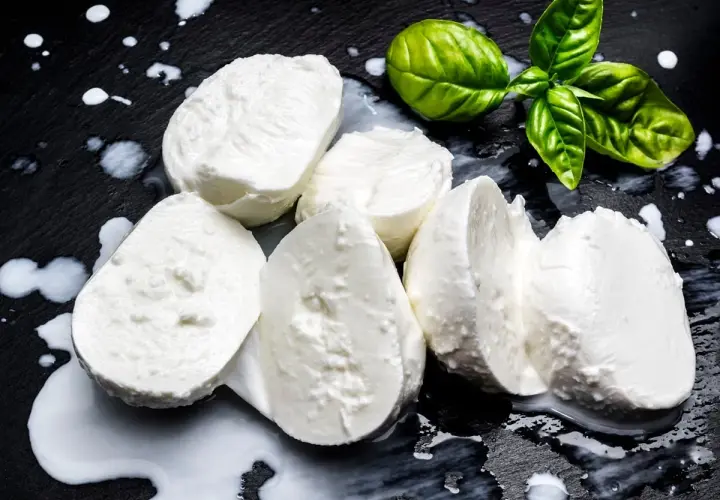 Świeża mozzarella na czarnym tle, z kilkoma listkami bazylii. Czy mozzarella jest kaloryczna? To zależy od jej rodzaju, ale zazwyczaj jest umiarkowanie kaloryczna.