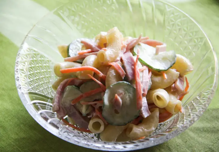 A close-up of a crystal bowl filled with creamy Japanese macaroni salad, featuring elbow macaroni, shredded carrots, sliced cucumbers, and ham.