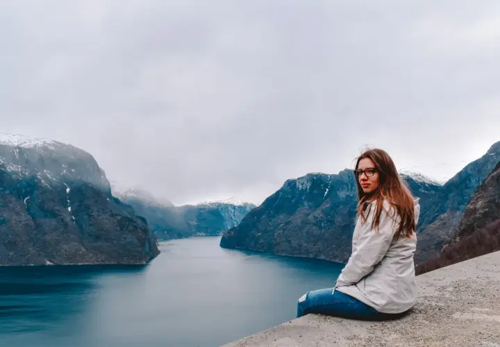 Kobieta siedzi na skarpie z widokiem na norweski fiord i ośnieżone góry.