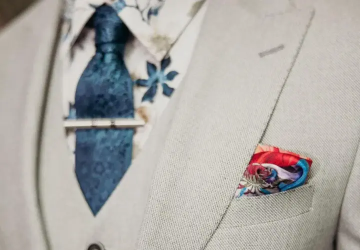 Elegancki mężczyzna w jasnej marynarce, z niebieskim wzorzystym krawatem i spinką do krawata jak nosić. W kieszeni kolorowa poszetka.