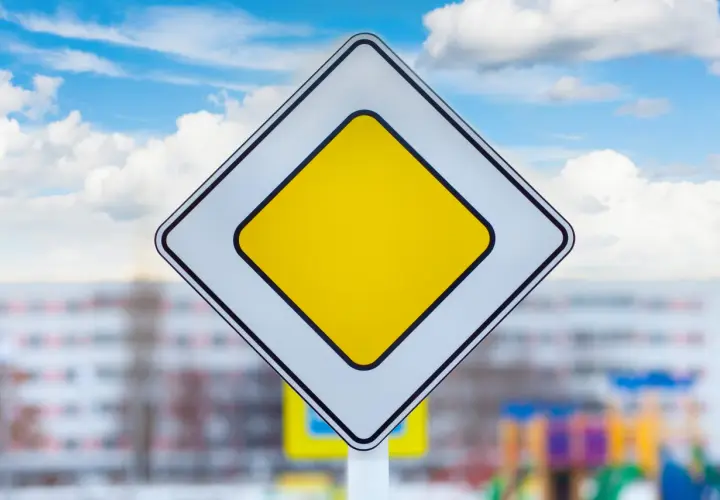 Znak droga z pierwszeństwem. Na tle błękitnego nieba z chmurami, biały romb z żółtym środkiem.