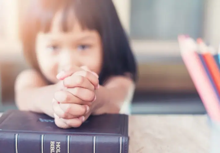 Dziecko z dłońmi złożonymi do modlitwy nad Biblią. Obok kolorowe kredki, symbol nauki dekalogu 10 przykazań.
