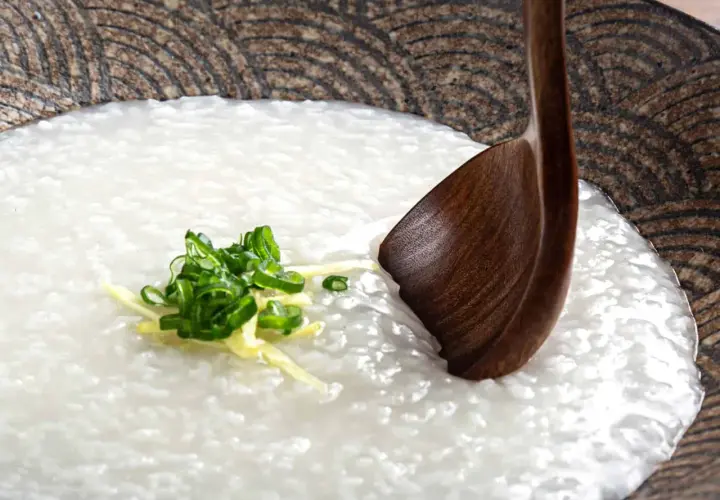 A comforting bowl of Japanese rice porridge (okayu) garnished with fresh scallions and ginger, ready to be enjoyed with a wooden spoon.
