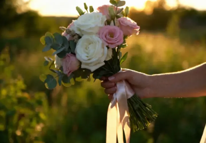 Dłoń panny młodej trzyma bukiet róż i eukaliptusa, ozdobiony satynową wstążką. Różne rodzaje wstążek florystycznych dodają uroku.