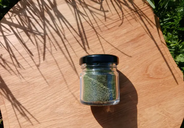 Un piccolo barattolo di vetro contiene polvere di ortica verde brillante, posato su un tagliere di legno con ombre di foglie.