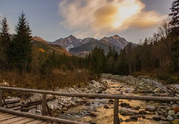 Jesienne Tatry w październiku. Drewniany mostek nad kamienistym potokiem, w tle góry i lasy.