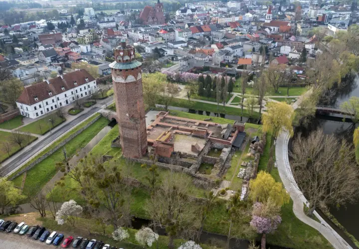 Ruiny zamku krzyżackiego w Brodnicy z charakterystyczną wieżą, otoczone zielenią i wodą, w tle rozciąga się miasto.