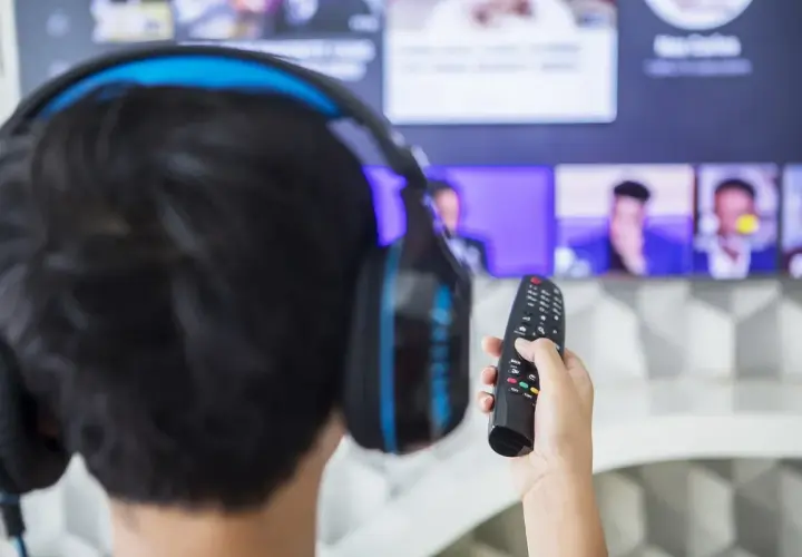 Chłopak w słuchawkach z pilotem w ręku, gotowy do oglądania na telewizorze Samsung. Dowiedz się, jak podłączyć słuchawki do Samsunga, by cieszyć się dźwiękiem.