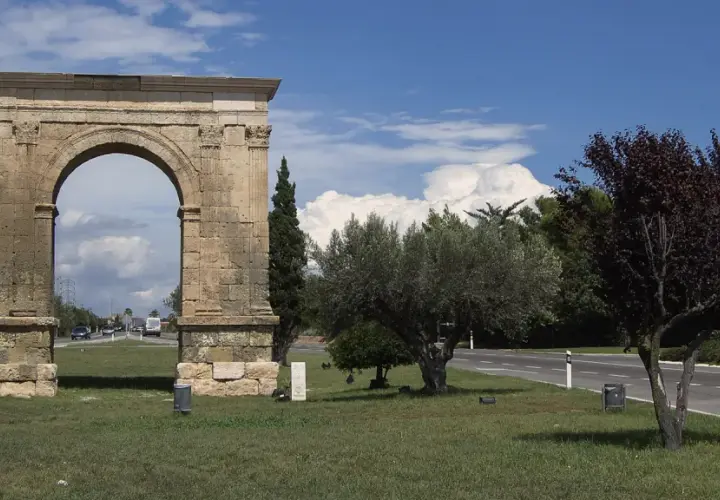 Arco romano de piedra, parte de una antigua roda de bara, flanqueado por árboles y palmeras bajo un cielo azul con nubes.