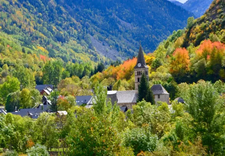 Pueblo en el **Valle de Arán** rodeado de árboles con hojas de otoño en tonos verdes, amarillos y naranjas. Destaca una iglesia con campanario.