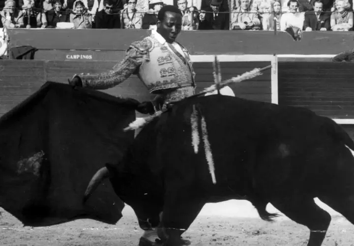 Un torero negro, con traje de luces, se enfrenta a un toro bravo en la arena. El público observa desde las gradas.