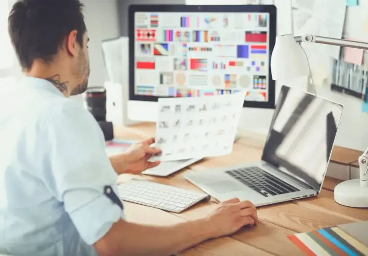 Mężczyzna, jako ux designer, analizuje wydruki przy biurku z komputerem i laptopem.