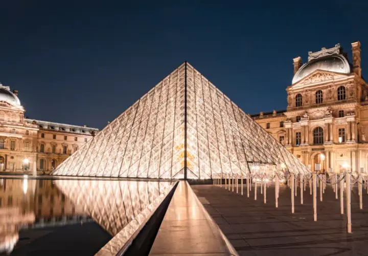 Piramide Luwru nocą z odbiciem w wodzie i oświetlonymi budynkami muzeum.