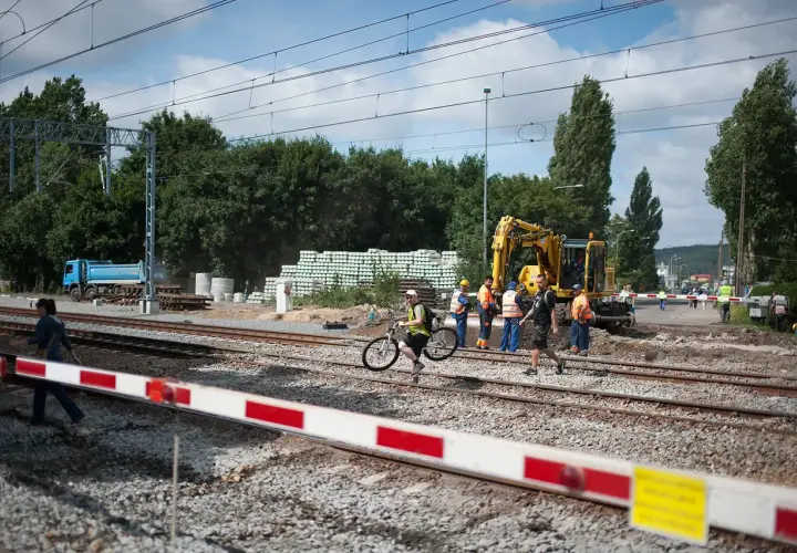 Pracownicy budowlani i dróżnik przejazdowy nadzorują prace przy torach. Koparka i ciężarówka są na miejscu.