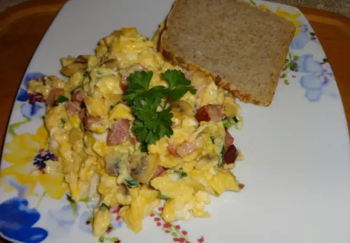 Pyszna jajecznica z boczkiem, pieczarkami i szczypiorkiem, udekorowana natką pietruszki, podana z kromką chleba.
