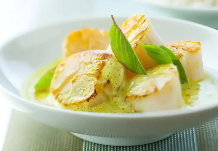 Délicieuses noix de saint-jacques poêlées, nappées d'une sauce onctueuse au curry et vin blanc, garnies de feuilles vertes.