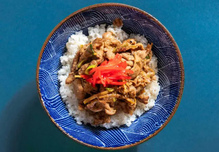 A bowl of fluffy white rice topped with savory pork shogayaki, garnished with bright red pickled ginger.