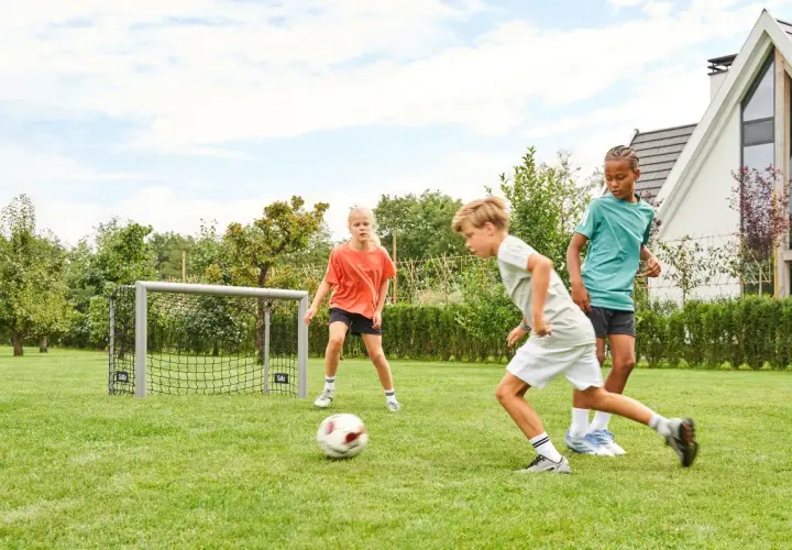 Drei Kinder üben im Garten Fußball. Ein Junge dribbelt, während ein Mädchen und ein weiterer Junge zuschauen.