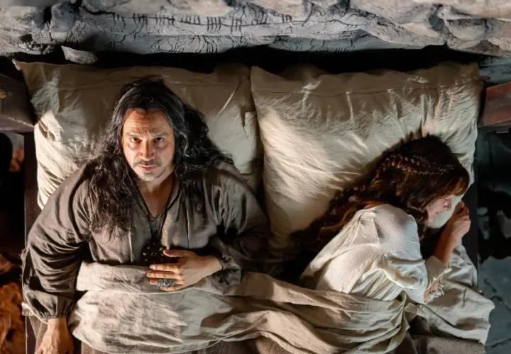 Un homme aux longs cheveux regarde la caméra, allongé dans un lit à côté d'une femme endormie. Un air de mystère plane sur ce moment, rappelant les intrigues de Mordred dans Kaamelott.