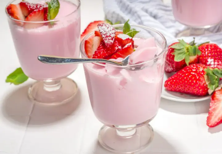 Różowa panna cotta z truskawkami, udekorowana listkami mięty i posypana cukrem pudrem. Obok świeże truskawki.