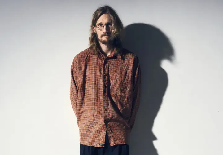 Marcus Berggren komiker, med sitt karaktäristiska hår och glasögon, står framför en vit vägg med en skugga som dansar bakom honom.