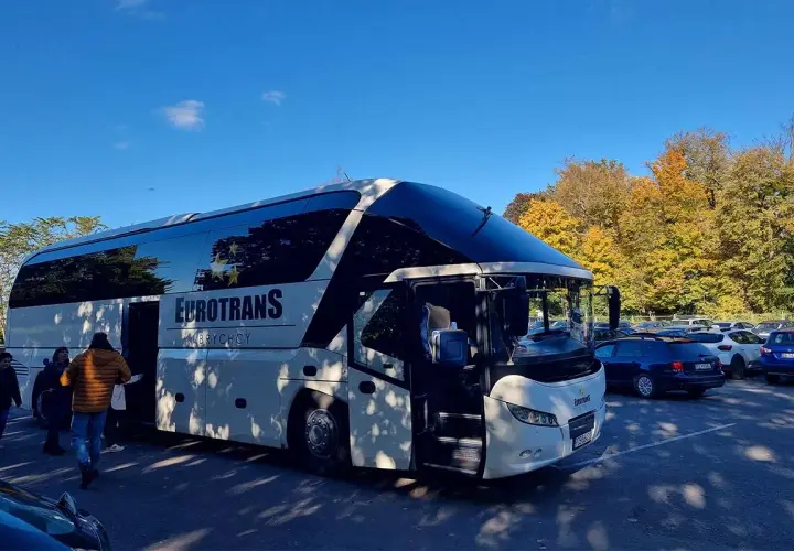 Biały autokar firmy Eurotrans z otwartymi drzwiami, zaparkowany na tle jesiennych drzew i innych samochodów.