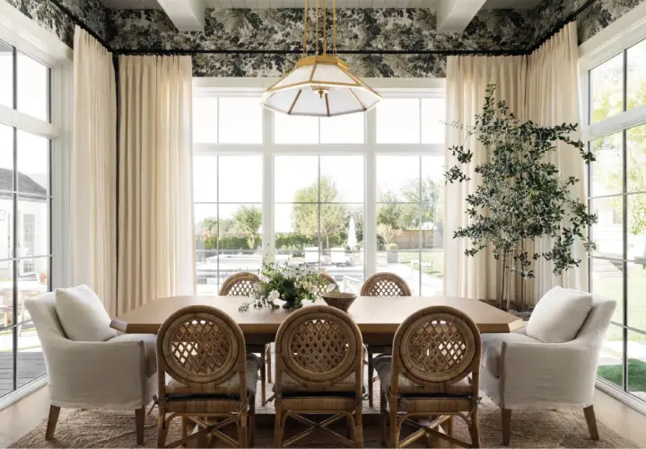 A bright dining room in a Studio McGee home features a large wooden table with rattan chairs, plush armchairs, and a floral wallpaper.