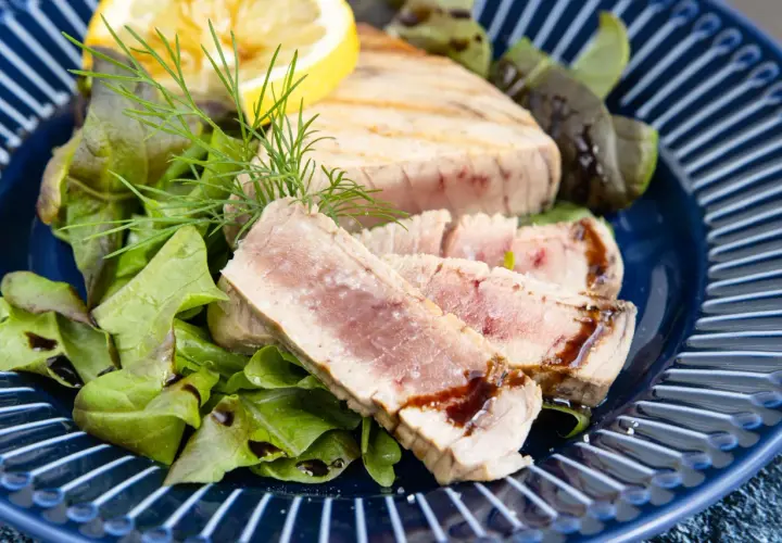 Saftiges Thunfisch Steak, medium gebraten, serviert mit frischem Salat, Dill und einer Zitronenspalte auf einem blauen Teller.