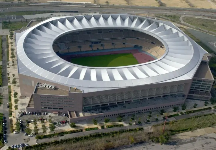 Imponujące, okrągłe **Estadio La Cartuja** z charakterystycznym, falującym dachem, otoczone zielenią i drogami.