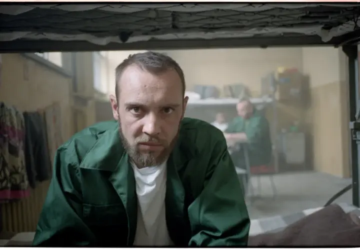 Mężczyzna w zielonej kurtce, jak z filmu "Jak zostałem gangsterem. Historia prawdziwa", patrzy wprost. W tle inni więźniowie.