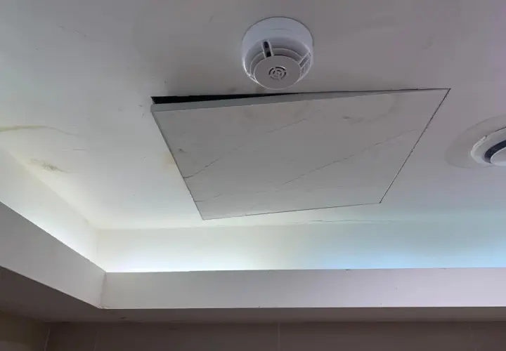 Un soffitto in cartongesso con una botola aperta, un rilevatore di fumo e macchie d'acqua. Il montaggio botola cartongesso è visibile.