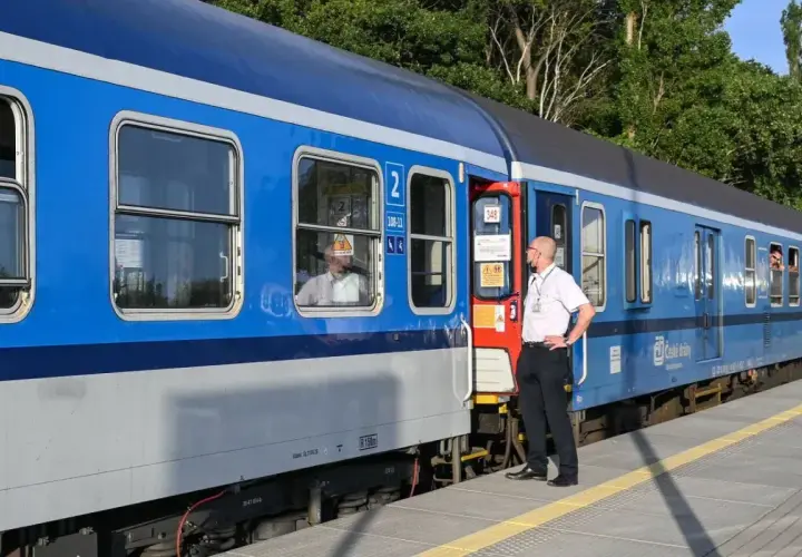 Pracownik kolei stoi przy drzwiach niebieskiego pociągu. Pasażerowie wyglądają przez okna. Czy można kupić bilet w pociągu?