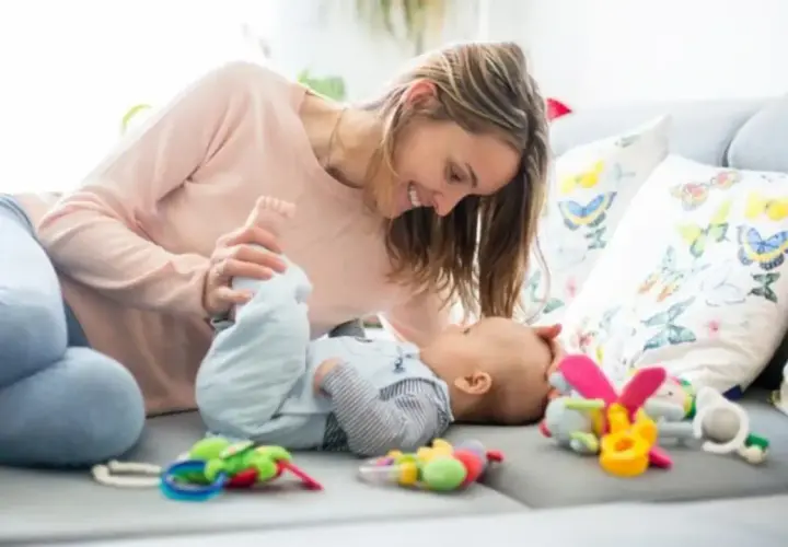 Mamá sonríe a su bebe de 2 meses mientras juegan en el sofá entre coloridos juguetes.