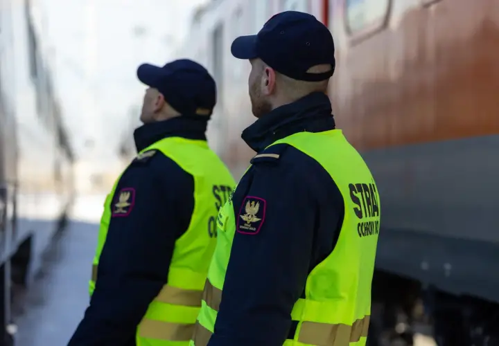 Dwóch funkcjonariuszy straży ochrony kolei w kamizelkach odblaskowych stoi przy pociągu, czuwając nad bezpieczeństwem.